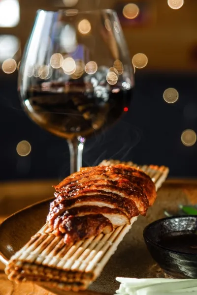 Detailaufnahme: Peking Ente mit Pfannkuchen, Frühlingszwiebeln, Gurke und Hoisin-Sauce – zarte Entenhaut und saftiges Fleisch auf hauchdünnem Pfannkuchen, Wein im Hintergrund